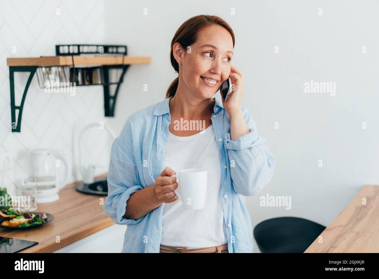 Jeune femme souriante attrayante avec une tasse de café dans la cuisine parlant au téléphone. Banque D'Images