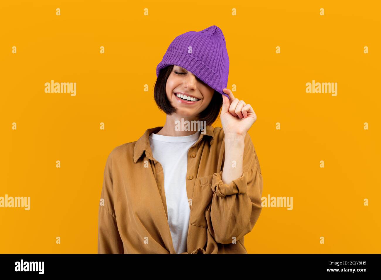 Bonne jeune femme tirant vers le bas chapeau violet tendance, fermant un oeil et souriant large, fond orange Banque D'Images