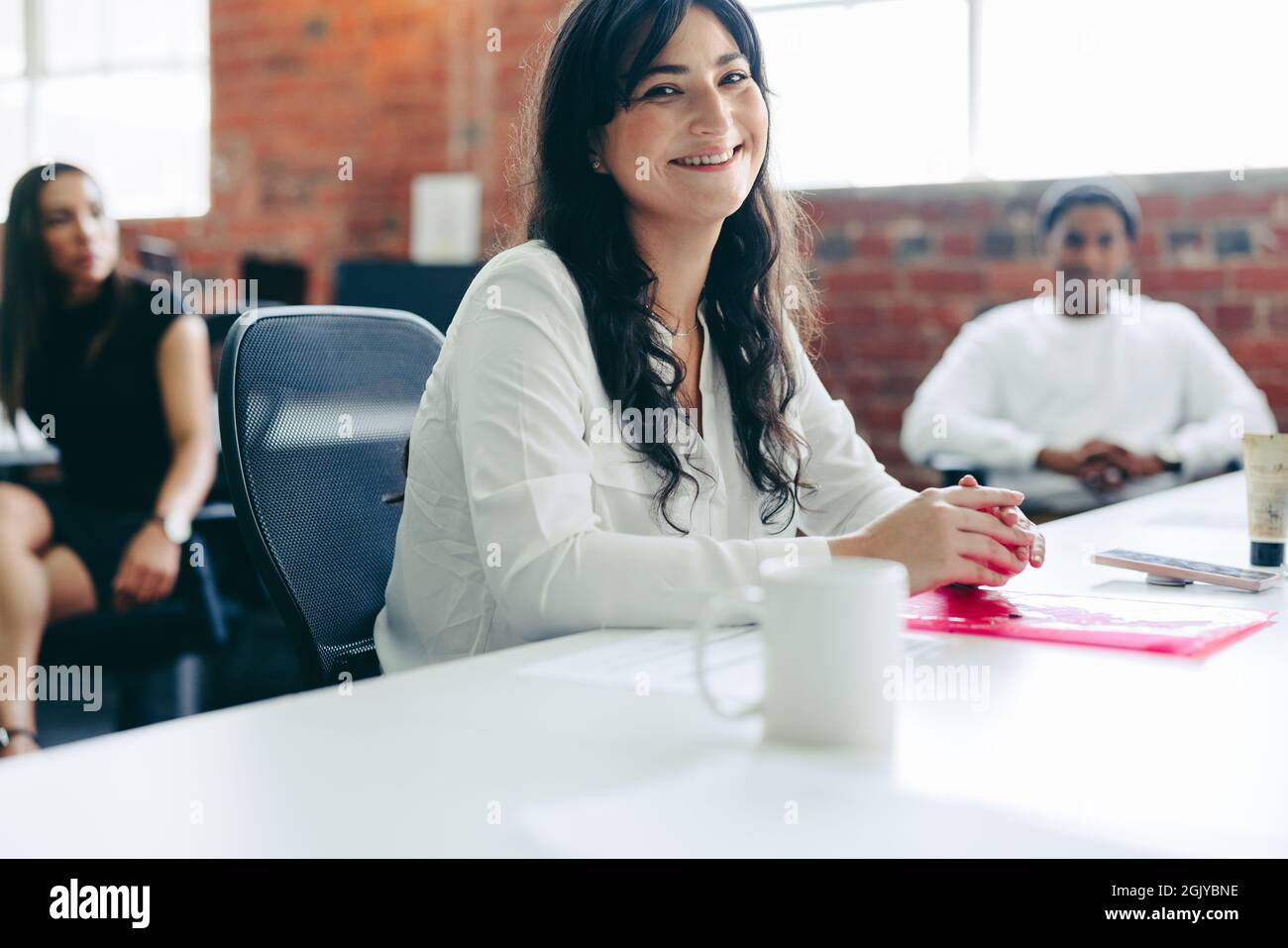 Femme d'affaires créative souriant dans un bureau avec ses collègues en arrière-plan. Bonne jeune femme d'affaires regardant l'appareil photo pendant Banque D'Images