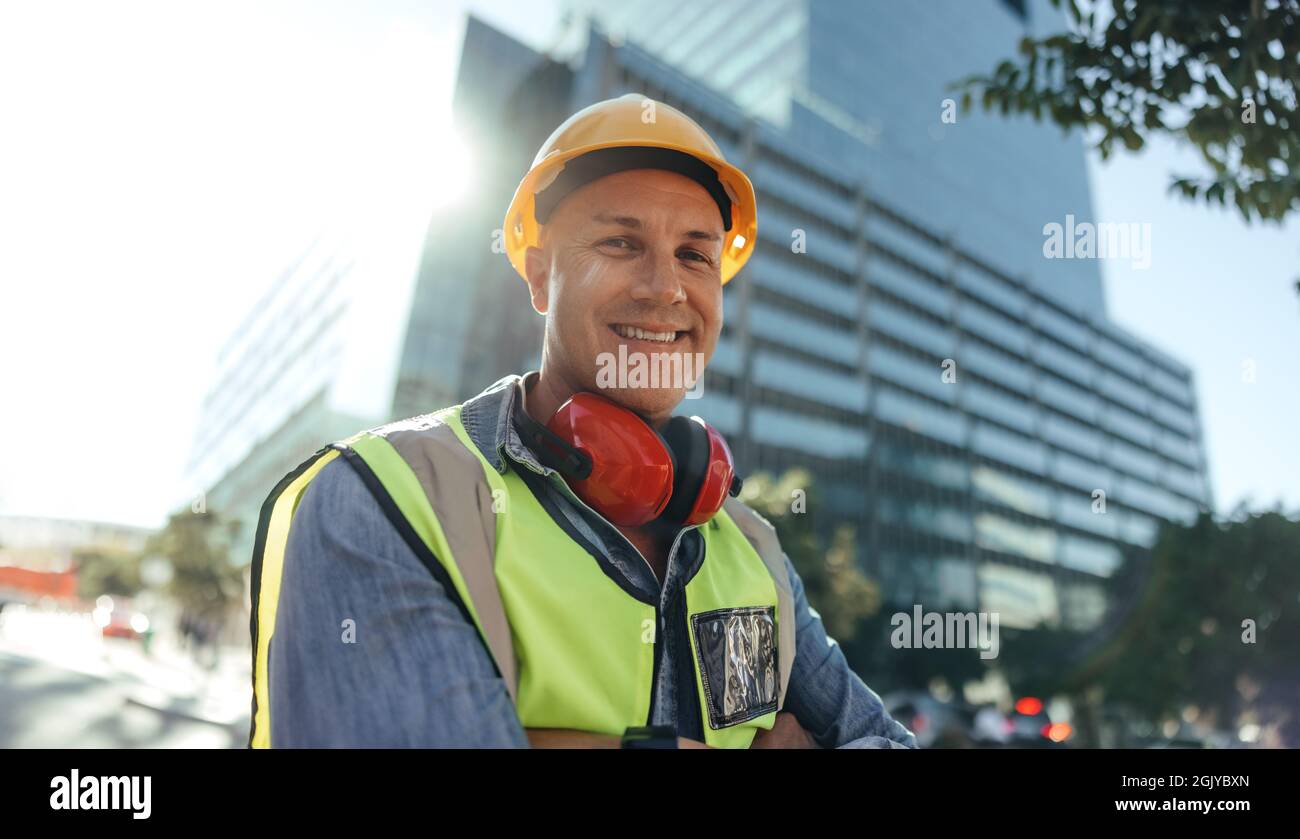 Joyeux ouvrier de construction souriant à la caméra tout en se tenant debout avec ses bras croisés dans la ville. Employé de col bleu pour adulte de taille moyenne debout devant Banque D'Images Joyeux ouvrier de construction souriant à la caméra tout en se tenant debout avec ses bras croisés dans la ville. Employé de col bleu pour adulte de taille moyenne debout devant Banque D'Images