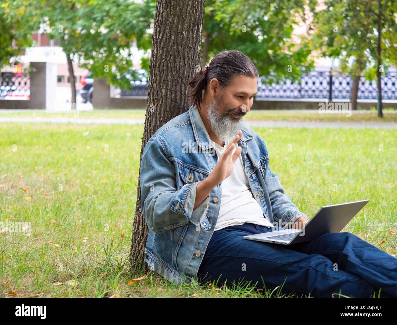 Photo d'un homme d'âge moyen pendant un appel vidéo en ligne. Il est assis sur l'herbe dans le parc de la ville avec un ordinateur portable. Il agite la main - Bonjour. Banque D'Images