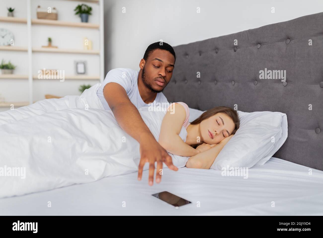 Homme afro jaloux vérifiant le téléphone mobile de sa copine dormanteuse dans le lit à la maison. Concept de problèmes de mariage Banque D'Images