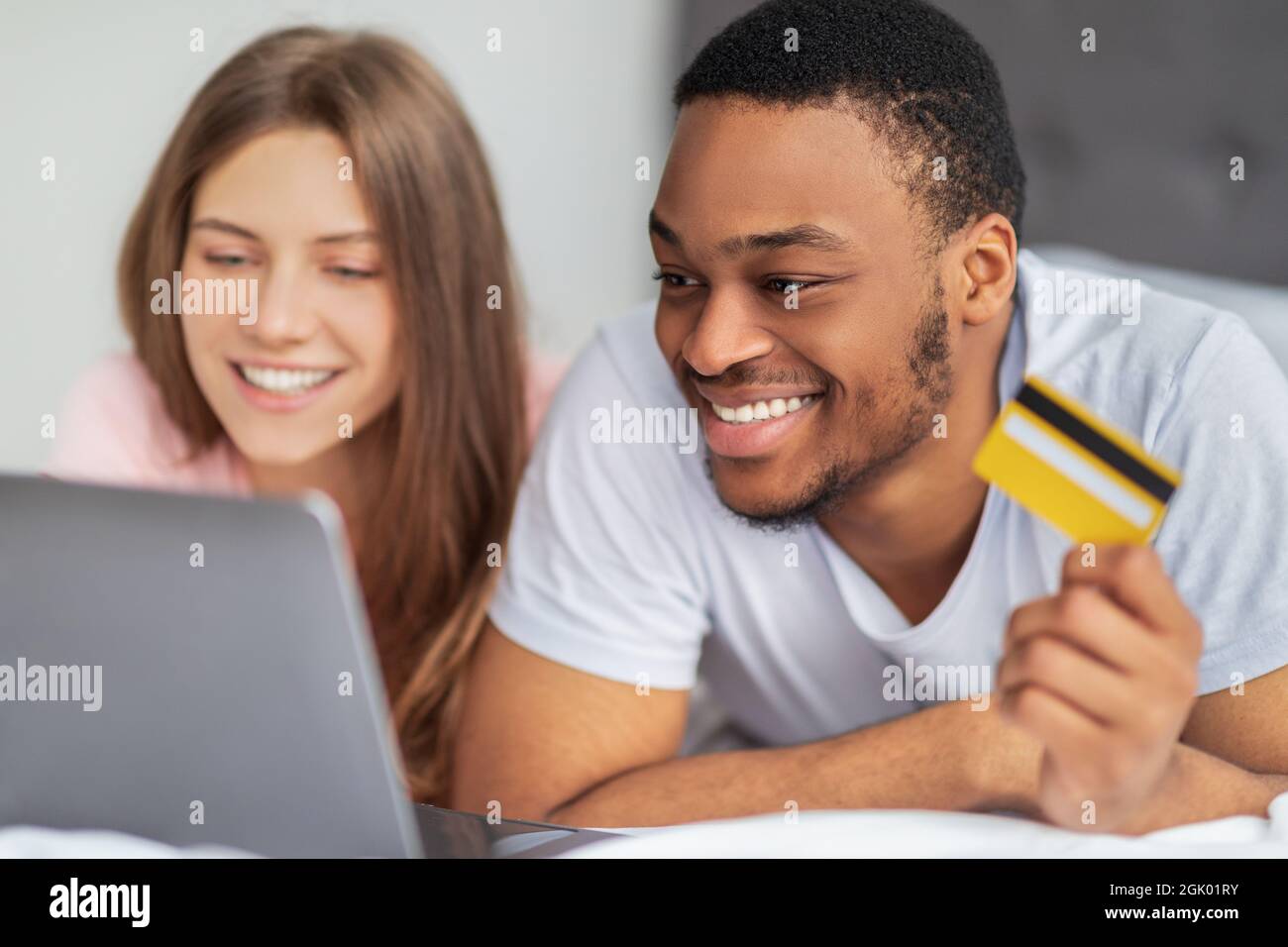 Portrait d'un couple multiracial heureux avec carte de crédit et ordinateur portable couché sur le lit, shopping en ligne de la maison Banque D'Images