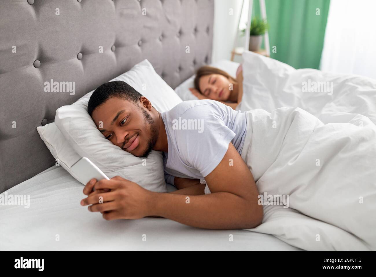 Jeune homme noir allongé sur le lit et utilisant le téléphone portable pour envoyer des SMS à son amant pendant que sa femme est endormi. Infidélité à la relation Banque D'Images