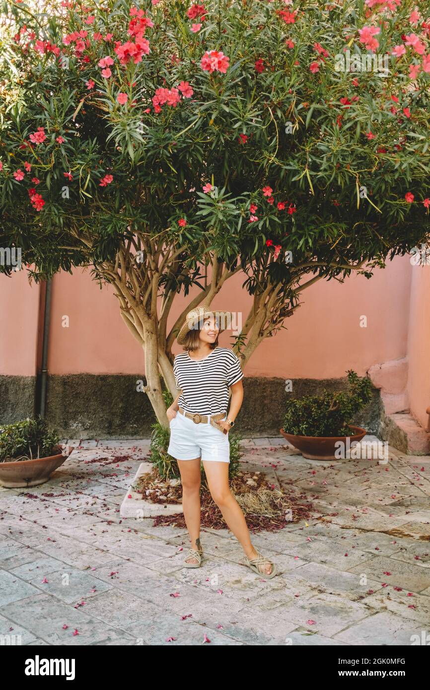Jolie femme touristique debout près de bougainvilliers arbre en fleurs dans l'île grecque - Corfou. Concept de déplacement. Tenue tendance avec chapeau de paille. Banque D'Images