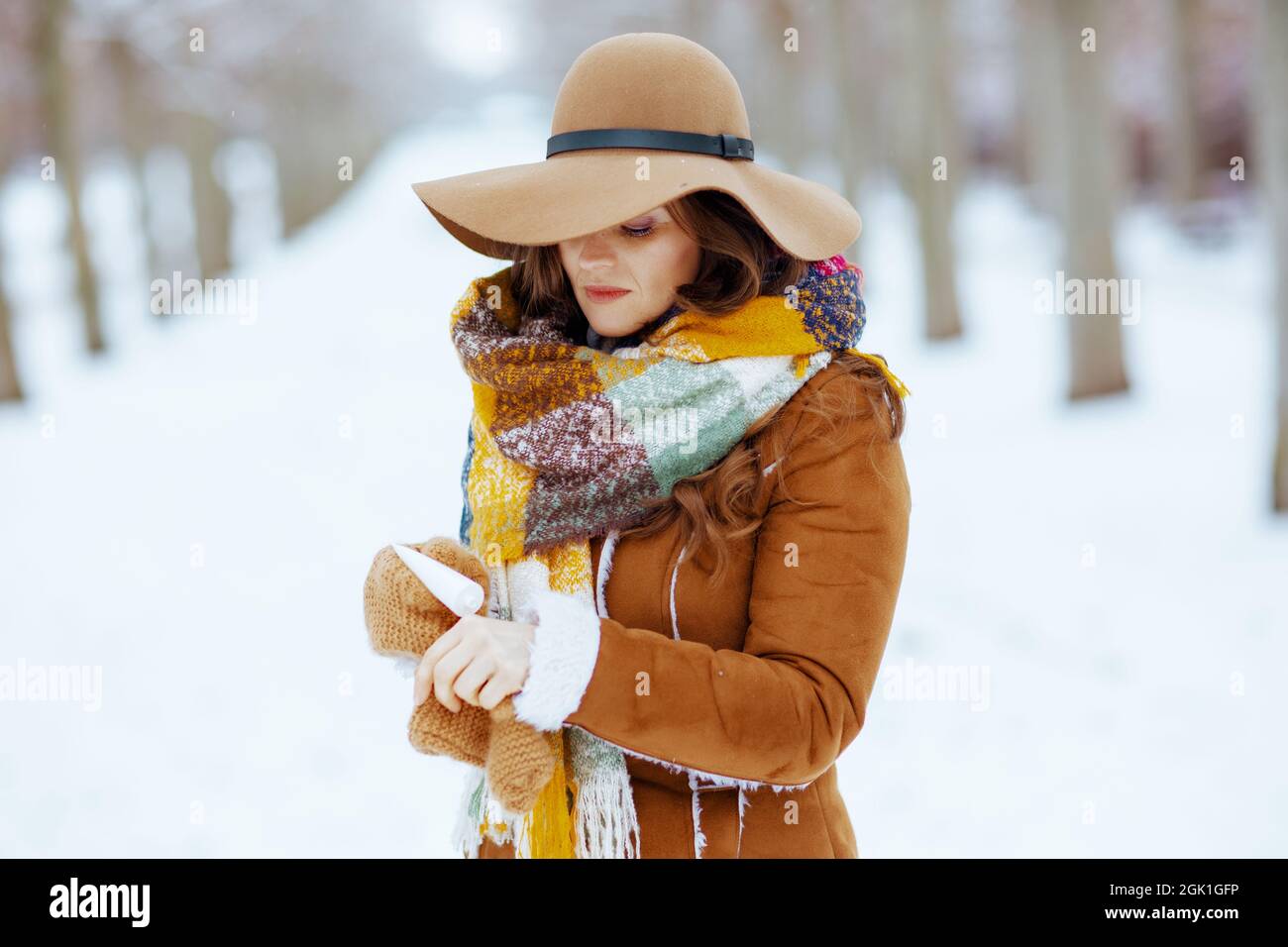élégante femme en chapeau marron et écharpe avec moufles en peau de mouton  en utilisant la crème pour les mains en plein air dans le parc de la ville  en hiver Photo