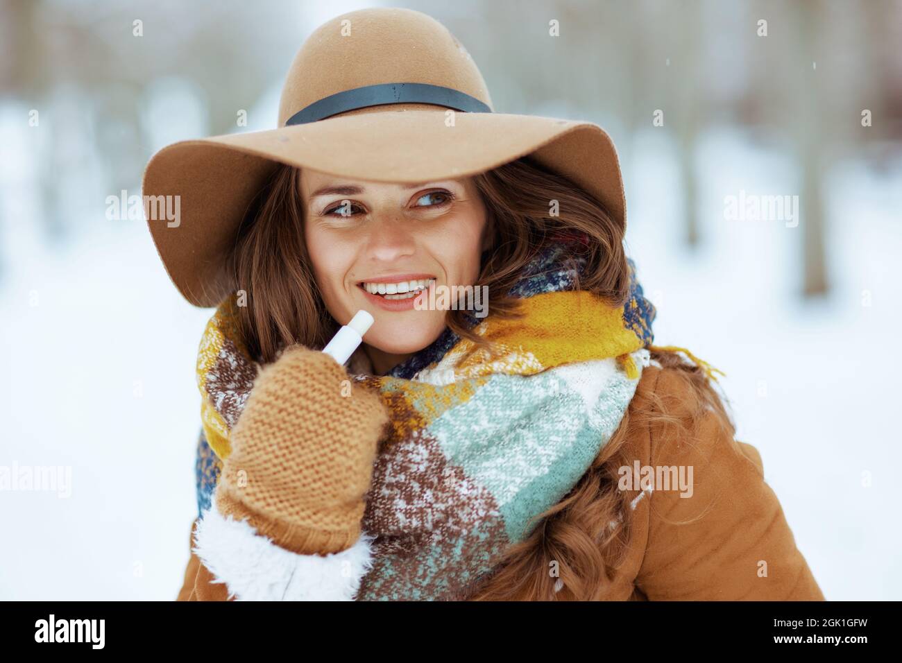 bonne femme élégante en chapeau marron et écharpe avec moufles et rouge à  lèvres hygiénique en peau de mouton en plein air dans le parc de la ville  en hiver Photo Stock -