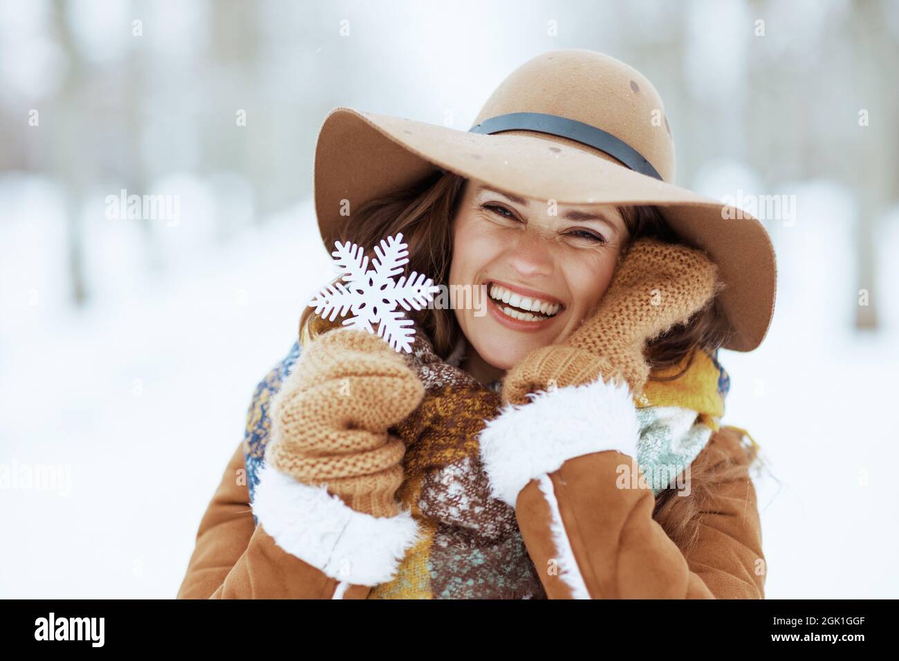 Portrait d'une femme élégante et souriante de 40 ans en chapeau marron et  écharpe avec moufles et flocons de neige en peau de mouton à l'extérieur du  parc de la ville en