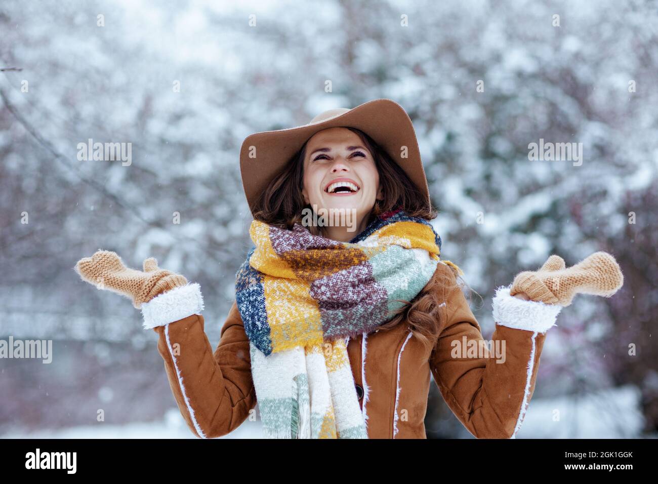 femme élégante et souriante de 40 ans en chapeau marron et écharpe avec  moufles en peau de mouton qui attrape la neige en plein air dans le parc de  la ville en