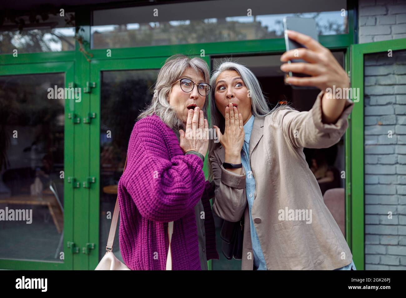 Drôles femmes senior prennent le selfie couvrant la bouche avec des palmiers sur la rue de la ville Banque D'Images