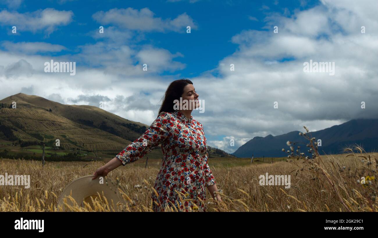 Bonne jeune femme en robe florale marchant au milieu d'un champ de blé avec un chapeau à la main lors d'une journée nuageux avec ciel bleu et montagnes dans le backgro Banque D'Images