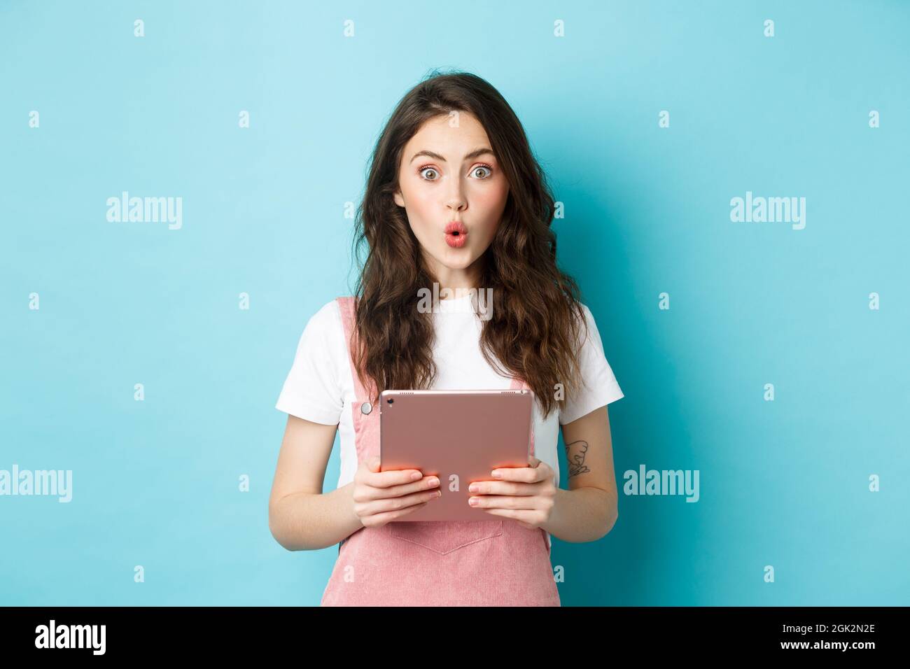 Impressionnée mignonne fille dire wow, rade l'excitation à l'appareil photo, tenant la tablette numérique sur la poitrine, debout dans les vêtements de printemps sur fond bleu Banque D'Images