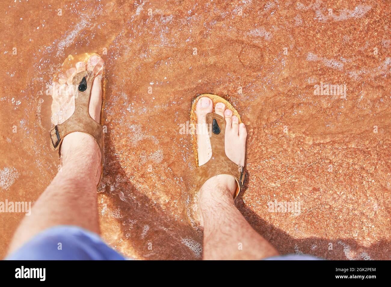 Pieds pour hommes dans des sandales sur la plage. L'homme se tient et regarde ses pieds dans le sable qui se lave par l'océan. Reposez-vous à l'hôtel. L'été. Banque D'Images