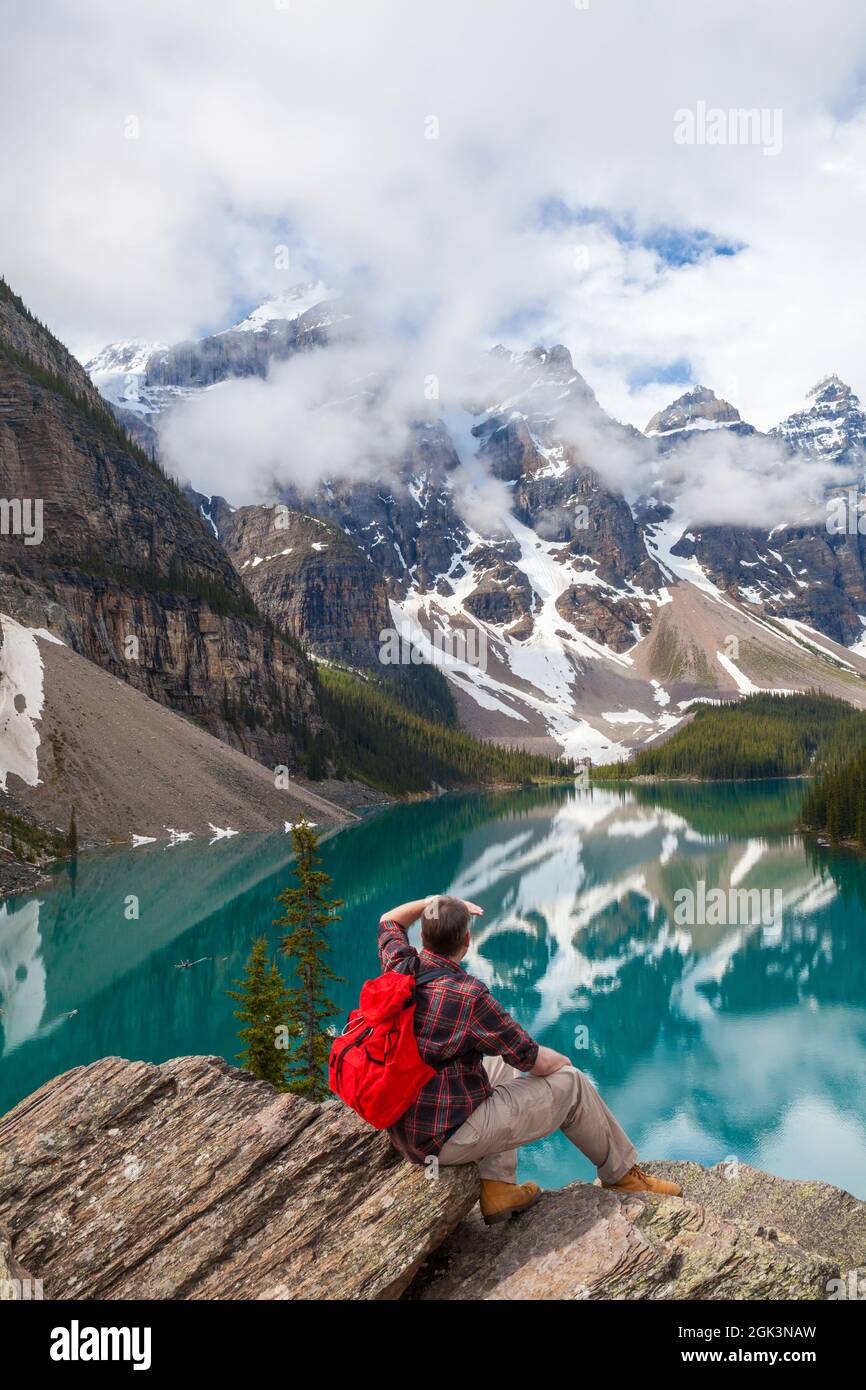 Homme de randonnée avec sac à dos à dos rouge assis sur un rocher surplombant le lac Moraine et regardant les sommets enneigés des montagnes Rocheuses, parc national Banff, Alabama Banque D'Images