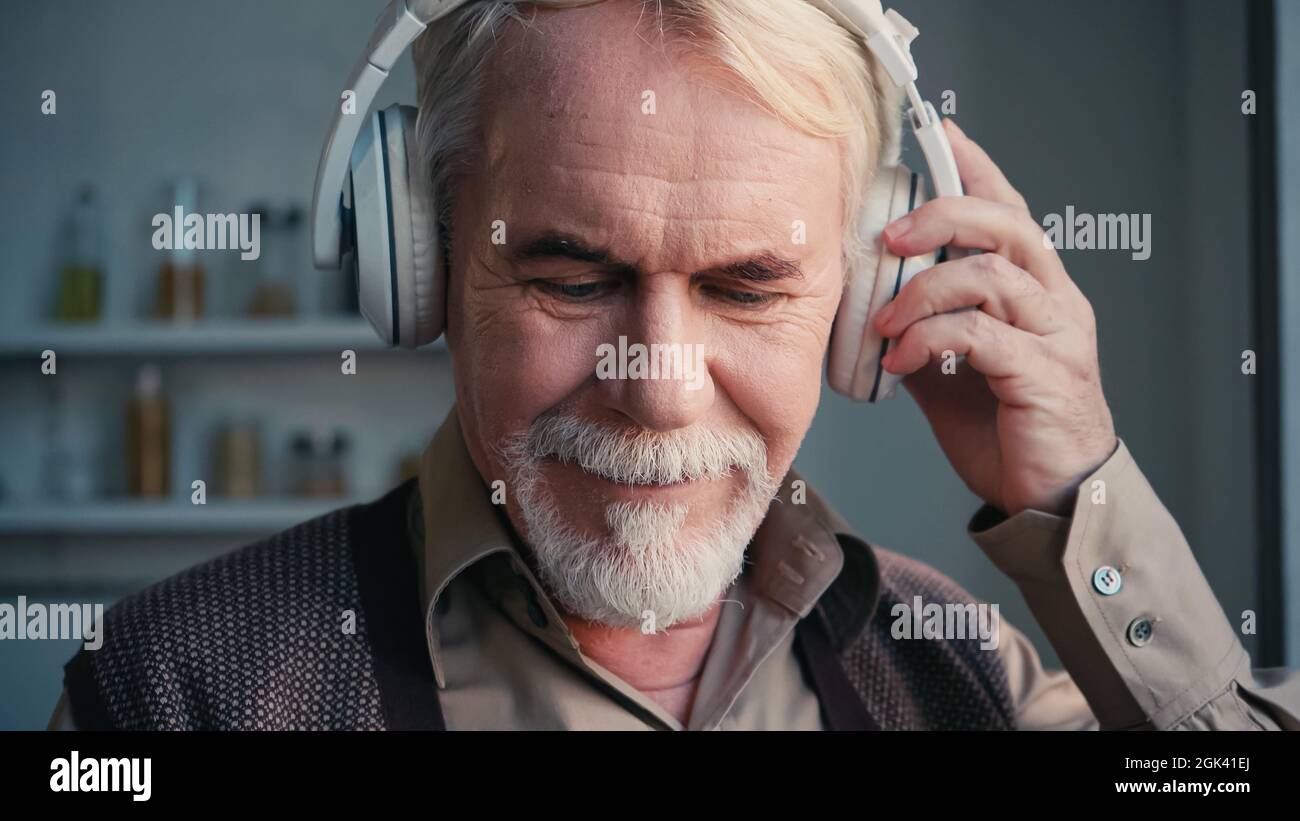 homme heureux et expérimenté qui règle les écouteurs sans fil tout en écoutant de la musique à la maison Banque D'Images