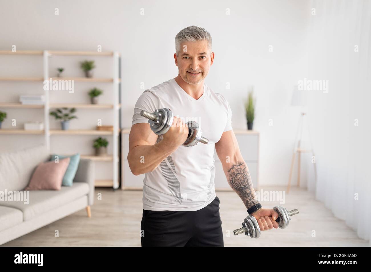 Entraînement sportif d'homme mature avec haltères à la maison, travail sur la force des bras, regarder et sourire à l'appareil photo, copier l'espace Banque D'Images