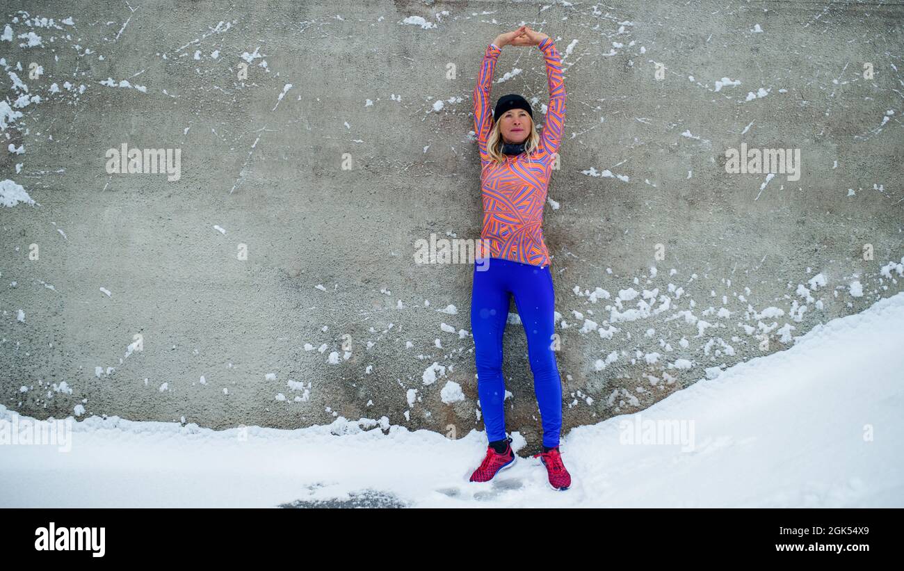 Femme senior active, debout contre un mur de béton à l'extérieur en hiver enneigé, s'étirant. Banque D'Images