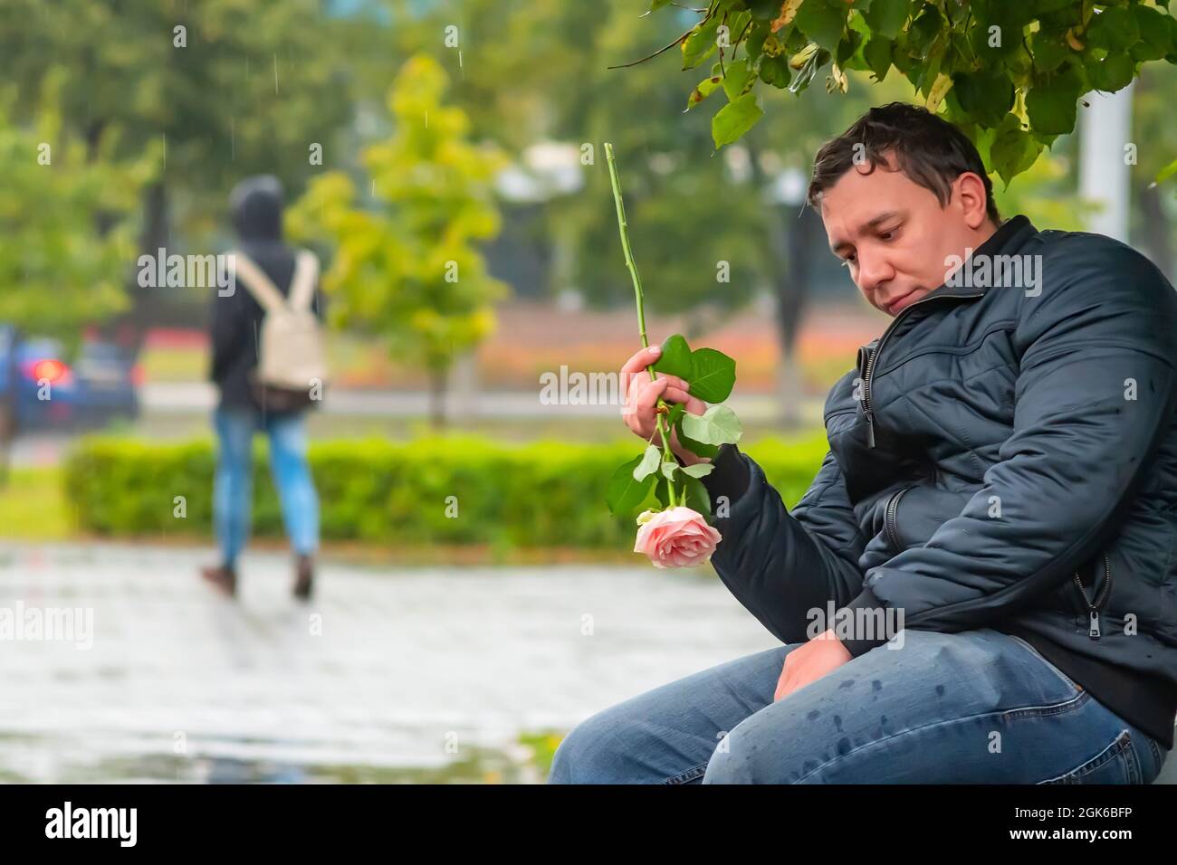 un triste homme regarde une fleur, un bourgeon d'une rose écarlate, assis, trempés dans la pluie sur un banc dans un parc de la ville Banque D'Images