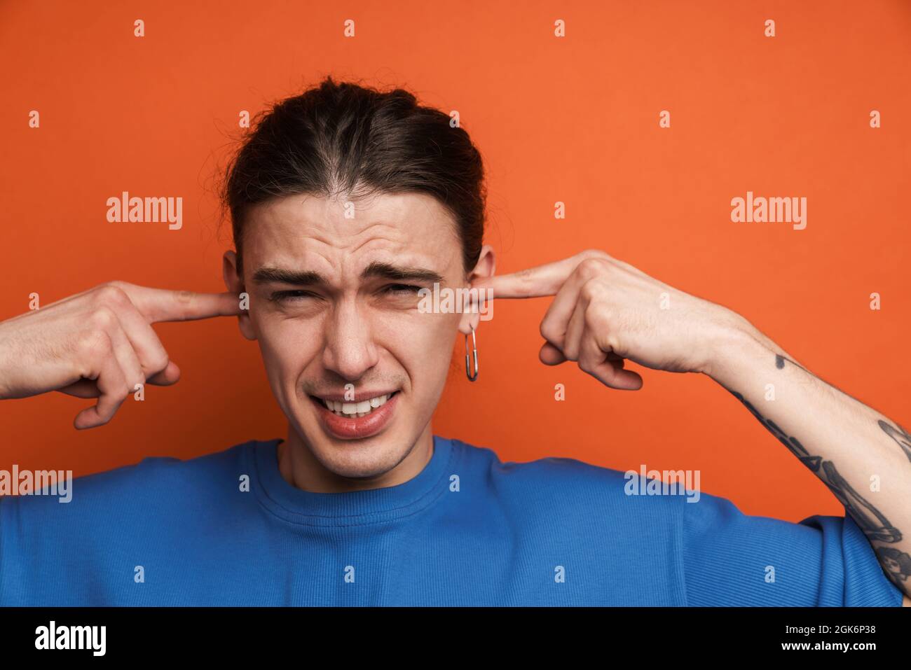 Jeune homme blanc avec des boucles d'oreilles frognant et bouchant ses oreilles isolées sur fond orange Banque D'Images