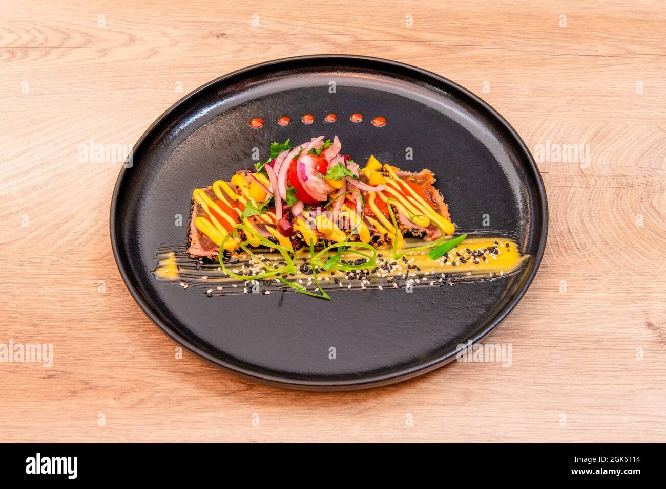 Assiette noire décorée de tatataki de thon rouge avec sauces, graines, oignons et grains de grenade sur fond de chêne Banque D'Images