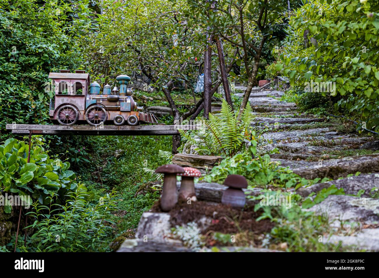 Une locomotive à vapeur miniature et des figurines de champignons en céramique décorent un chemin de jardin à Moghegno, encadré par des fougères et des arbres fruitiers. Circolo della Maggia, Tessin, Suisse. Banque D'Images