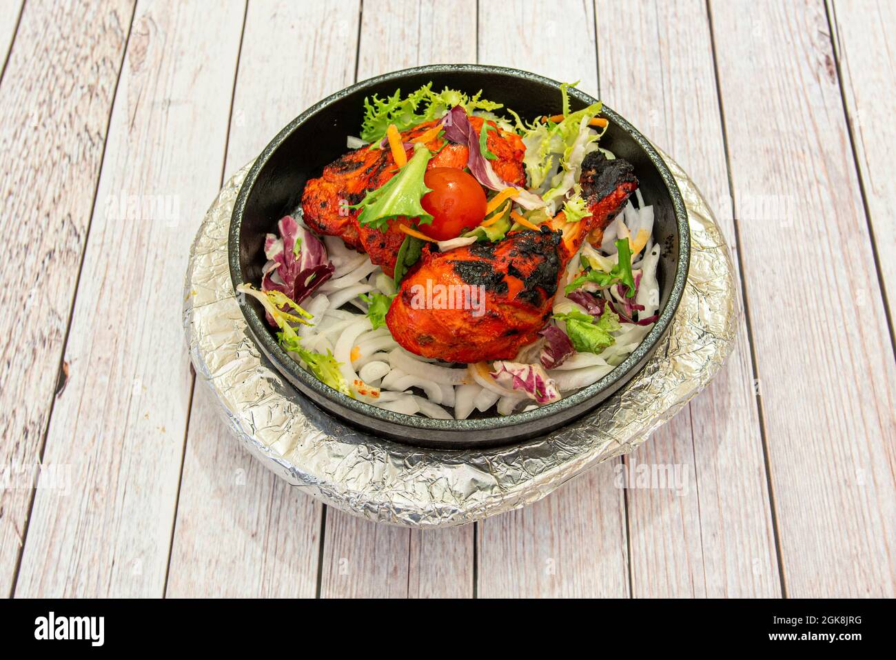 Bol en fonte chaud avec poulet tandoori rouge braisé garni de laitue, d'oignons et de tomates cerises sur table blanche Banque D'Images