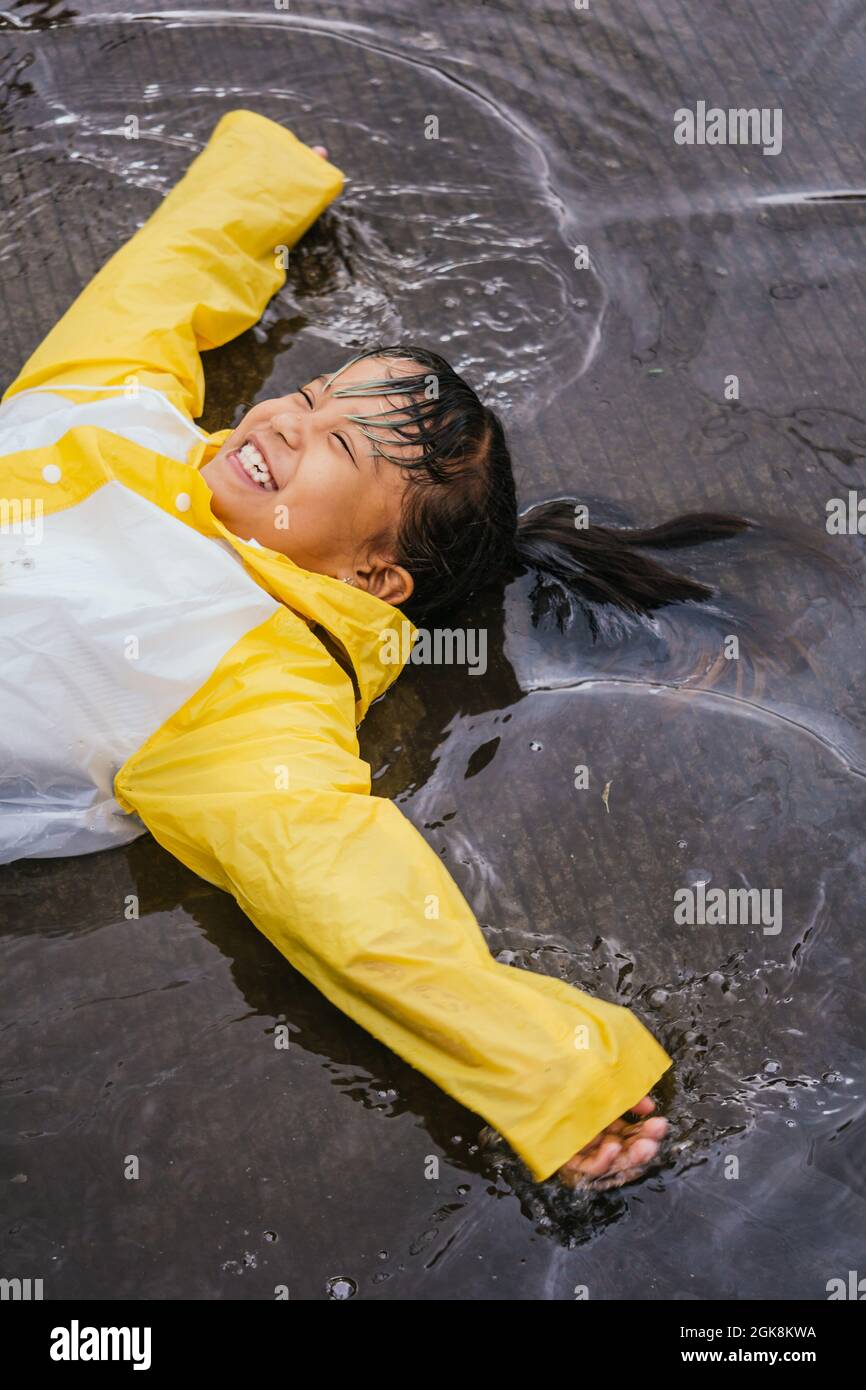 Grand angle de contenu asiatique Kid dans des bottes en caoutchouc et slicker couché dans la flaque ondulée le jour de la pluie Banque D'Images