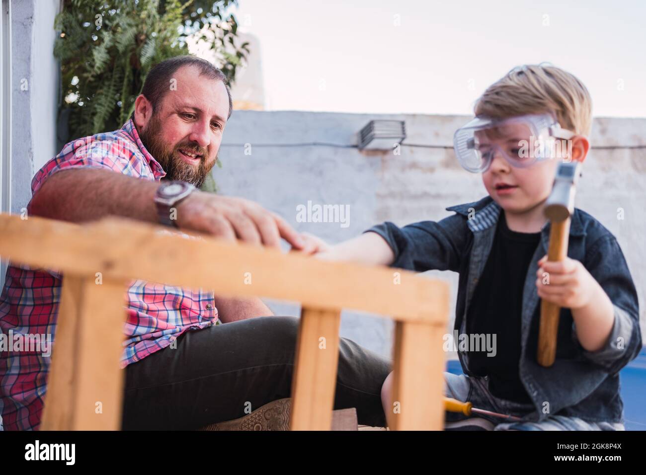 Vue latérale d'un papa hipster aidant son sur des lunettes de protection avec un marteau tout en faisant des travaux de bois Banque D'Images