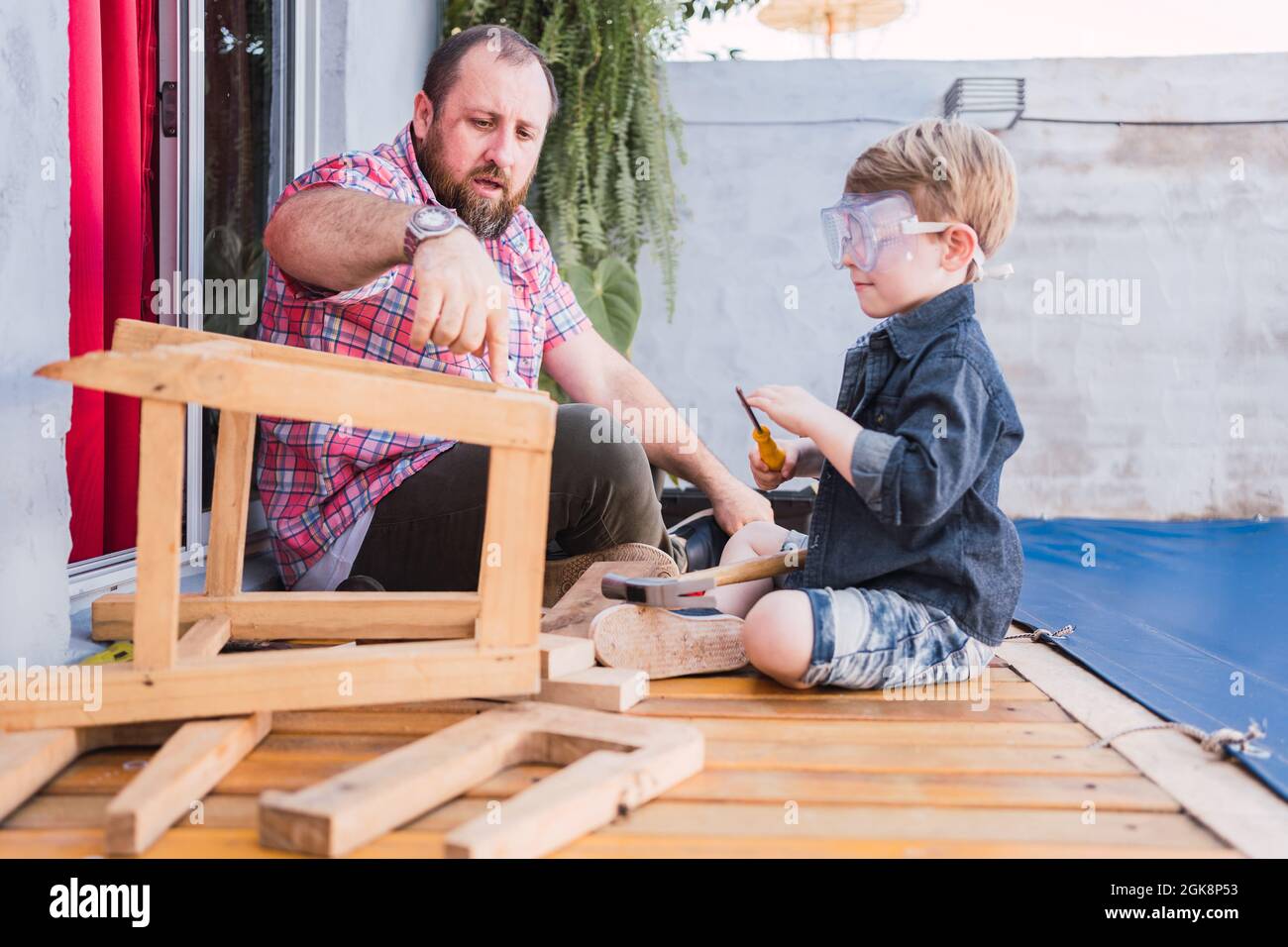 Père mûr expliquant à son fils dans des lunettes de protection comment conduire la vis dans le bloc de bois tout en étant assis sur la promenade Banque D'Images