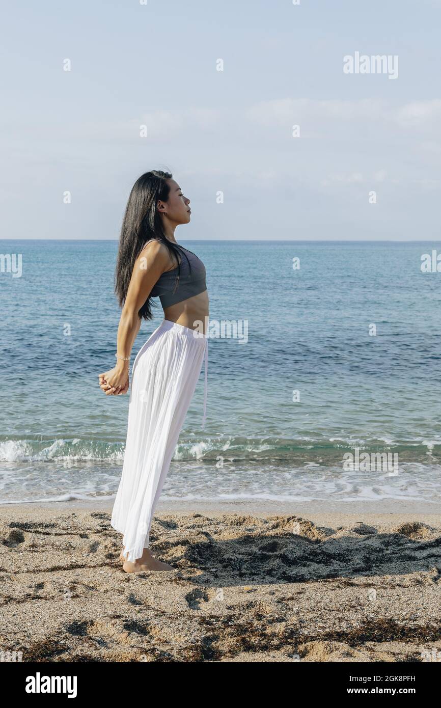 Vue latérale d'une femelle ethnique se pentant dans le dos tout en se tenant à Ashta Chandrasana pose pendant la pratique du yoga sur la côte sablonneuse contre l'océan Banque D'Images