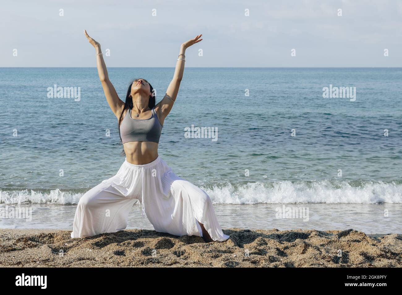 Jeune femme ethnique aux bras levés qui se squat sur la côte sablonneuse tout en pratiquant le yoga contre la mer en plein soleil Banque D'Images
