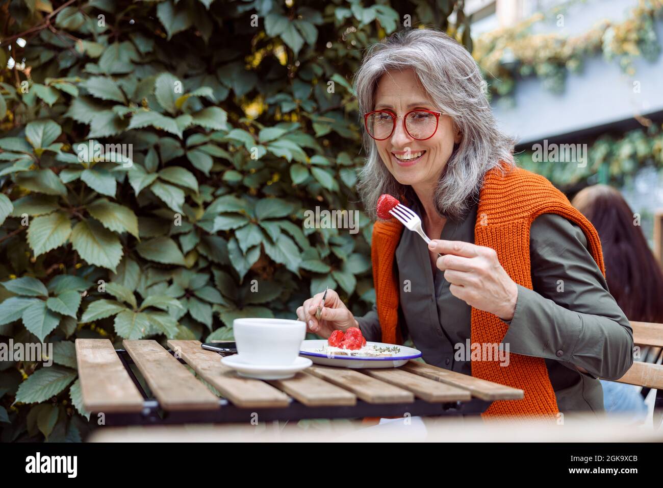 Une femme mûre souriante avec des verres mange des toasts à table sur la terrasse extérieure du café Banque D'Images