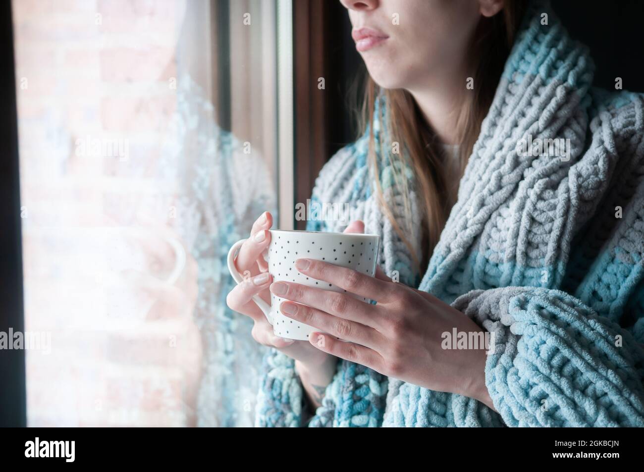 Jeune femme avec une couverture en laine en hiver, regardant par la fenêtre tenant une boisson chaude Banque D'Images