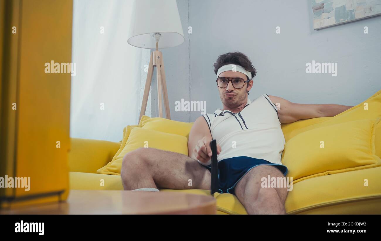 un jeune homme s'est ennuyé dans des lunettes et des vêtements de sport assis sur un canapé et à changer de chaîne Banque D'Images