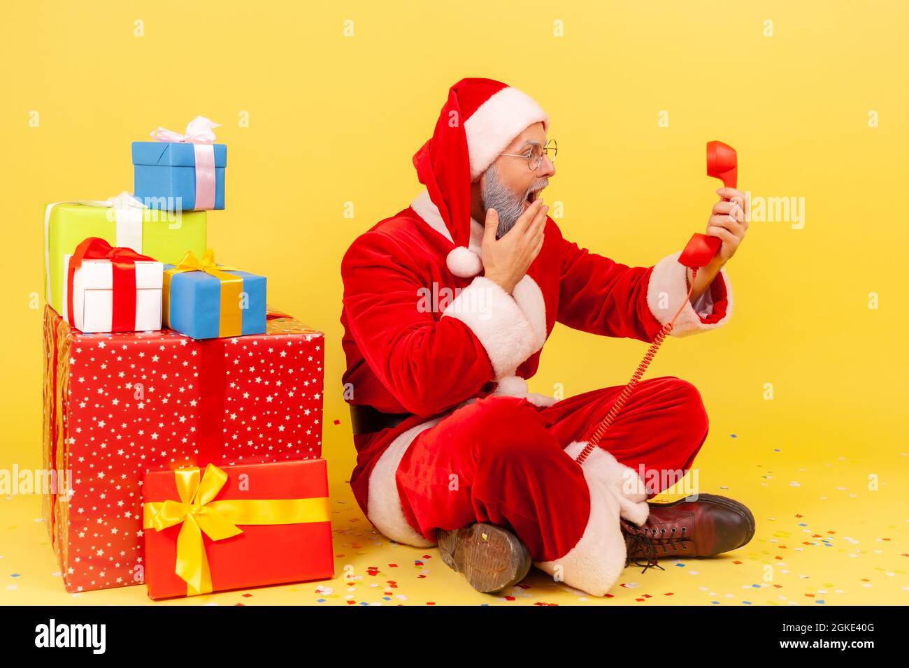 Un homme âgé surpris portant le costume du père noël assis sur le sol avec un téléphone rétro de la ligne terrestre, félicitations de Noël, couvrant la bouche avec la main. Po Banque D'Images