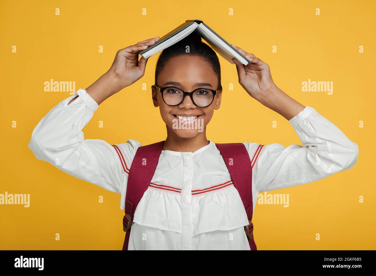Souriante et élégante, jeune femme afro-américaine en lunettes avec sac à dos tenant un livre sur la tête Banque D'Images
