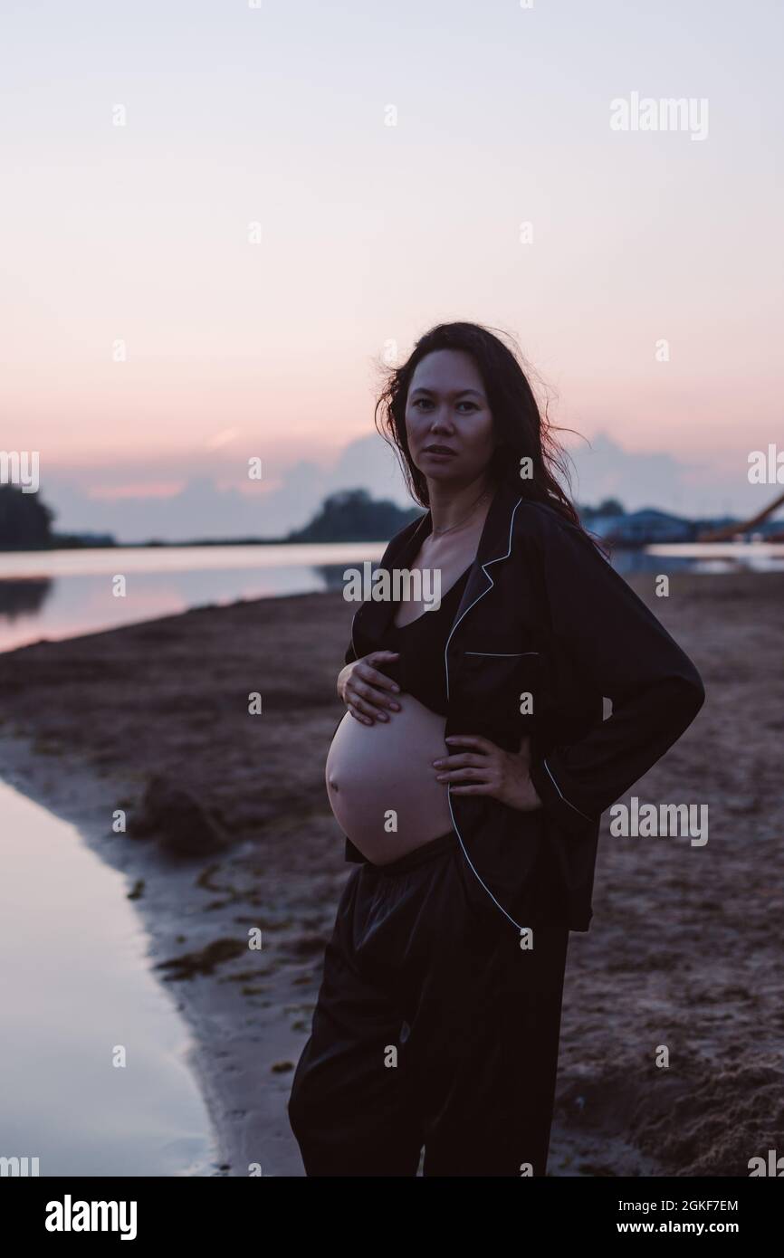 Portrait de femme enceinte sur la plage. Photo romantique de jeune brunette attrayante en pyjama sombre non boutonné sur la plage regardant l'appareil photo et ses cheveux Banque D'Images