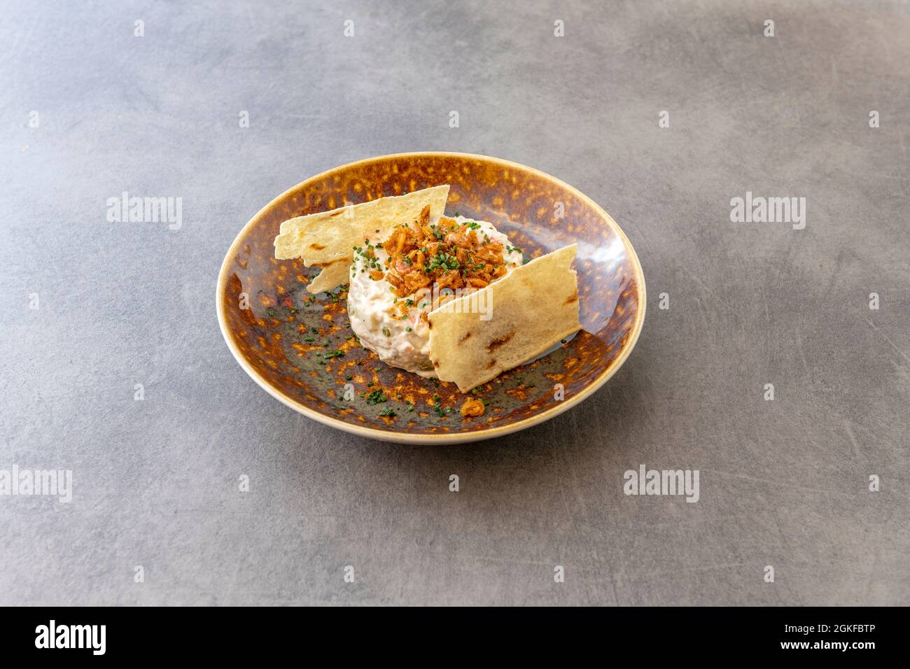 Tapa espagnole sophistiquée de salade russe surmontée de crevettes frites, ciboulette et accompagnée de pommes de terre croustillantes sur les côtés sur une assiette brune grillée Banque D'Images