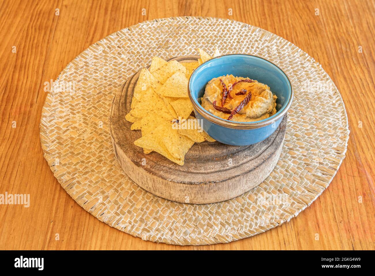 Petit bol bleu avec houmous de pois chiches, flocons de poivre séché et nachos à tremper sur un morceau de log et un trivet en osier Banque D'Images