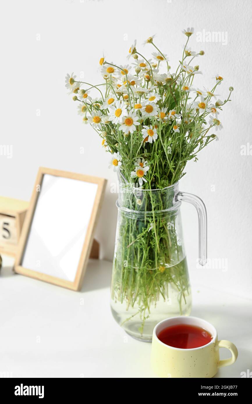 Vase avec des chamomiles et une tasse de thé sur une étagère près du mur lumineux Banque D'Images