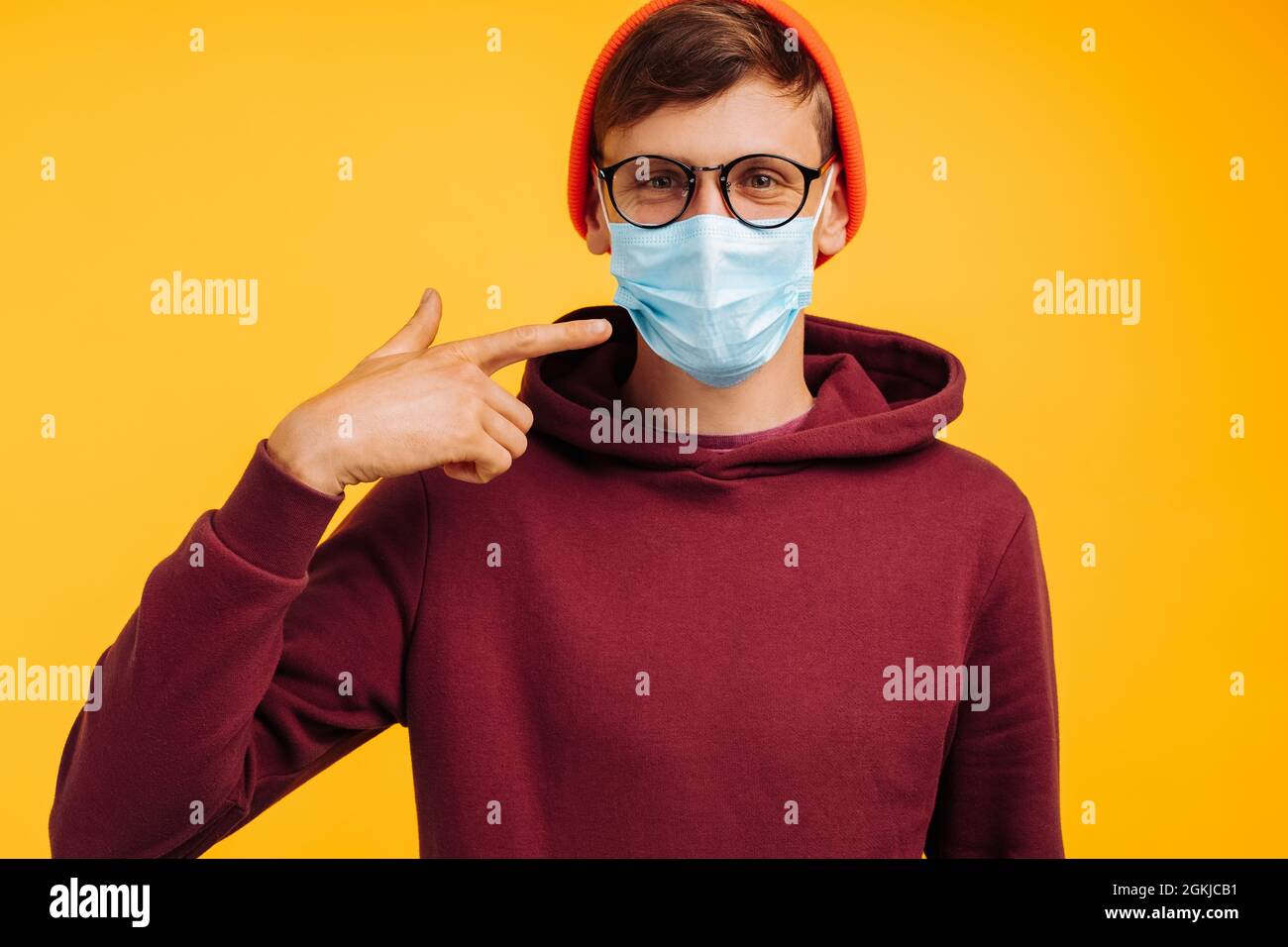 beau homme dans un masque de protection. en lunettes et un chapeau orange et un sweat-shirt rouge, souriant à l'appareil photo, sur fond jaune, heureux vacciné Banque D'Images