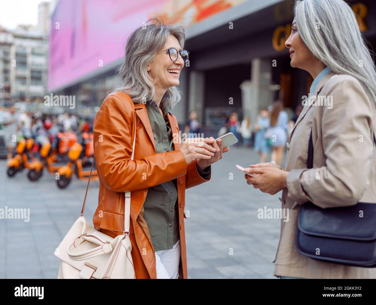 Souriante, femme d'âge mûr et femme asiatique avec téléphones mobiles modernes parlent dans la rue de la ville Banque D'Images