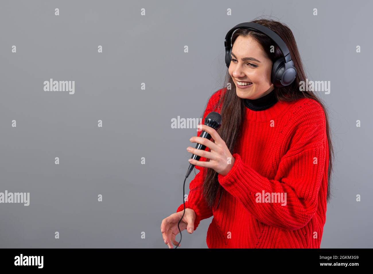 jeune, émotif brunette avec écouteurs et un microphone vêtu d'un chandail rouge chante karaoké, isolé sur gris Banque D'Images