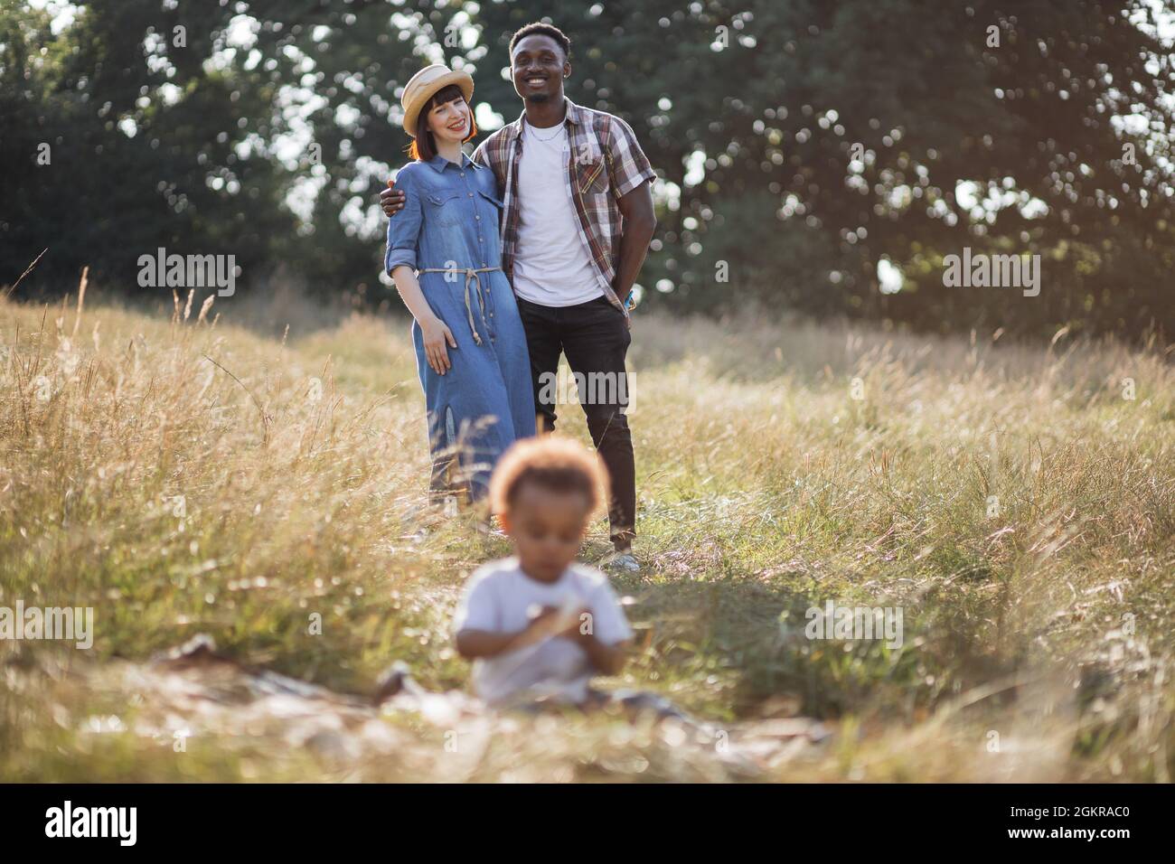 Flou de premier plan de petit garçon africain assis sur l'herbe et de jouer tandis que ses parents multiraciaux se tenant derrière, embrassant, souriant et regardant lui. Le bonheur de la parentalité. Banque D'Images