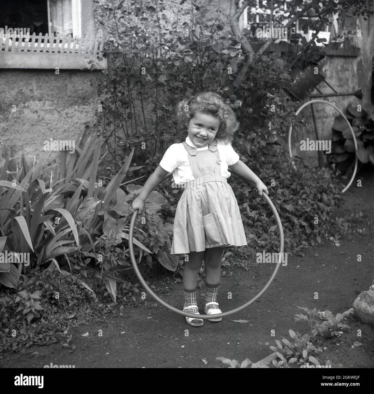 Années 1960, historique, à l'extérieur sur un chemin de jardin, une petite fille avec un hula hoop, un jouet hoop, autour de sa taille, Allemagne.Bien que le hula hoop ait été en existence et utilisé par les enfants pendant des centaines d'années, ce n'est qu'à la fin des années 1950, quand la version en plastique a été introduite, que la folie pour eux a pris le dessus.Le hula est devenu une mode mondiale, avec plus de 100 millions vendus en deux ans.Jusqu'à la fin des années 1970, au début des années 1980, les hula Hoops demeuraient un jouet populaire pour enfants. Banque D'Images