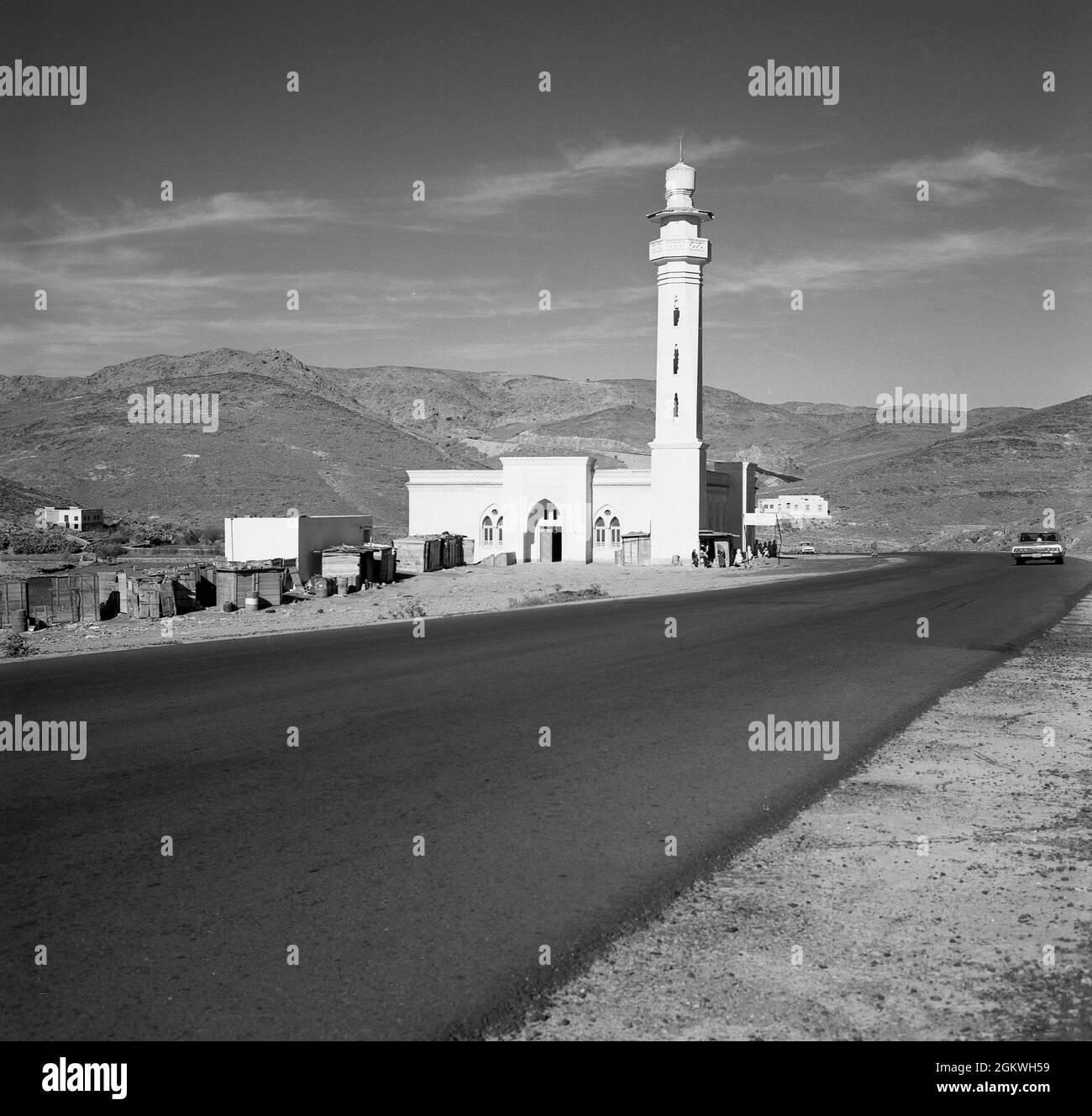 Années 1960, historique, dans une zone montagneuse aride, une vue sur une route tamac récemment mise en place, à côté d'elle un buiilding religieux arabe récemment construit, une mosquée, avec minaret ou tour, Arabie Saoudite. Banque D'Images