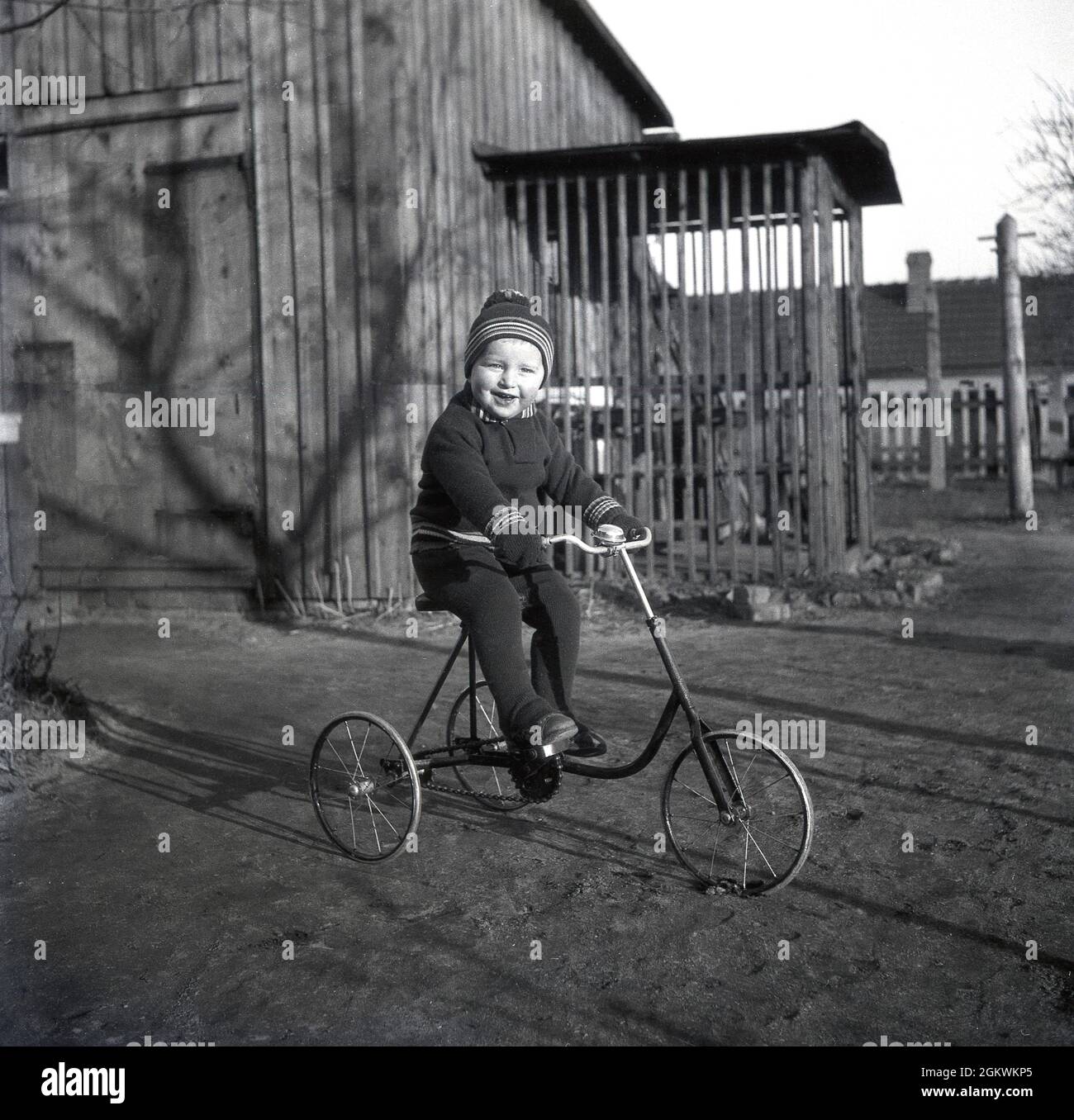 Dans les années 1950, ce petit garçon chignon, portant un ensemble de haut et de bas en coton côtelé, avec un chapeau à bulles assorti, assis à l'extérieur sur un tricycle en métal vintage, datant probablement des années 1930 et apparemment sans pneu en caoutchouc sur la roue avant. Banque D'Images