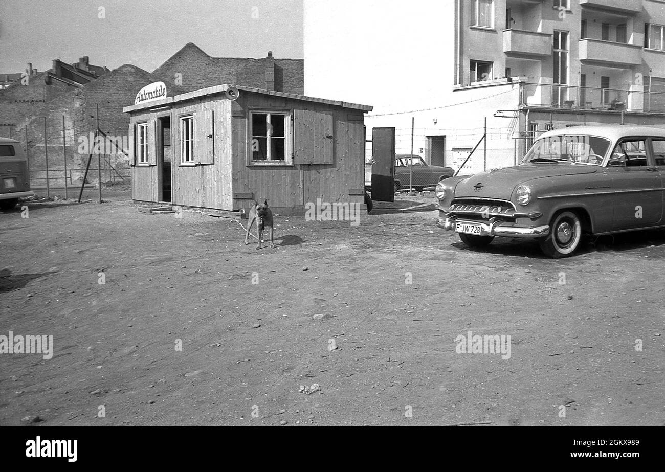 Années 1960, historique, parking d'occasion, Allemagne, situé à côté d'un bloc d'appartements, montrant une hutte en bois utilisée comme bureau situé sur une zone de gravier, avec une voiture de l'époque garée.Le panneau sur la hutte dit, "automobiles".Un chien de garde sur une chaîne se tient devant la cabane. Banque D'Images