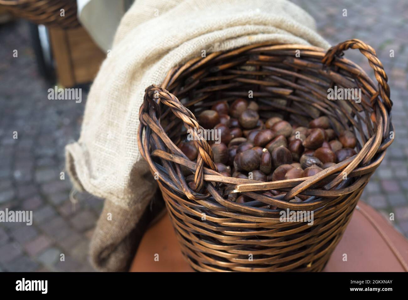 Panier en osier brun avec deux poignées pleines de châtaignes. Vente extérieure de produits agricoles. Couverture en lin léger et pavé de pavés Banque D'Images