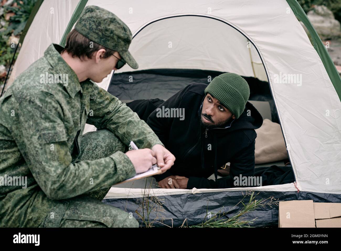 Soldat en uniforme de l'armée qui se croupe à la tente de l'homme noir tout en exécutant le compte de migrants Banque D'Images